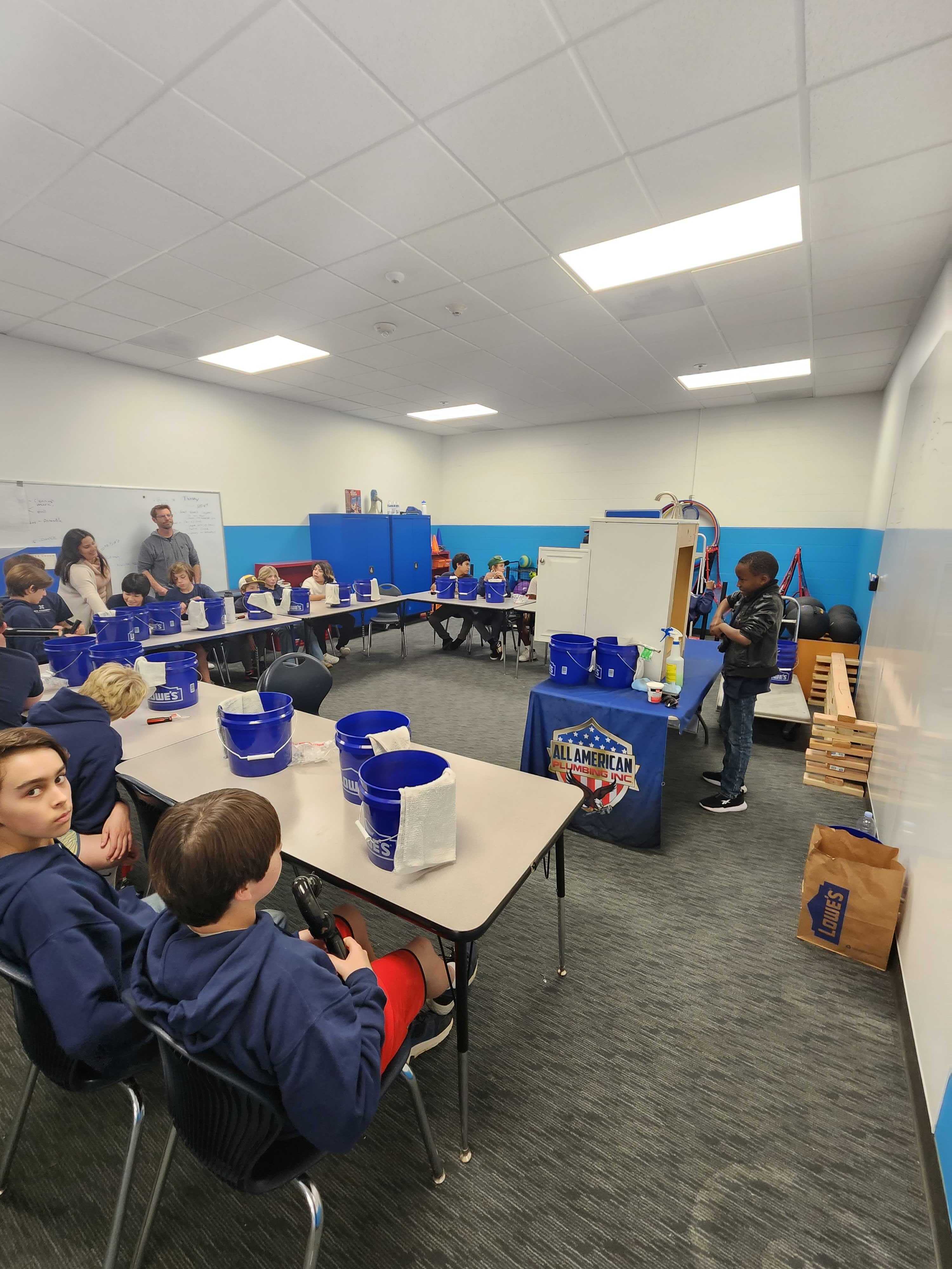 Children sitting around tables with blue Lowe's buckets listening to a young boy standing near a table covered with an All American Plumbing Inc. cloth in a classroom.