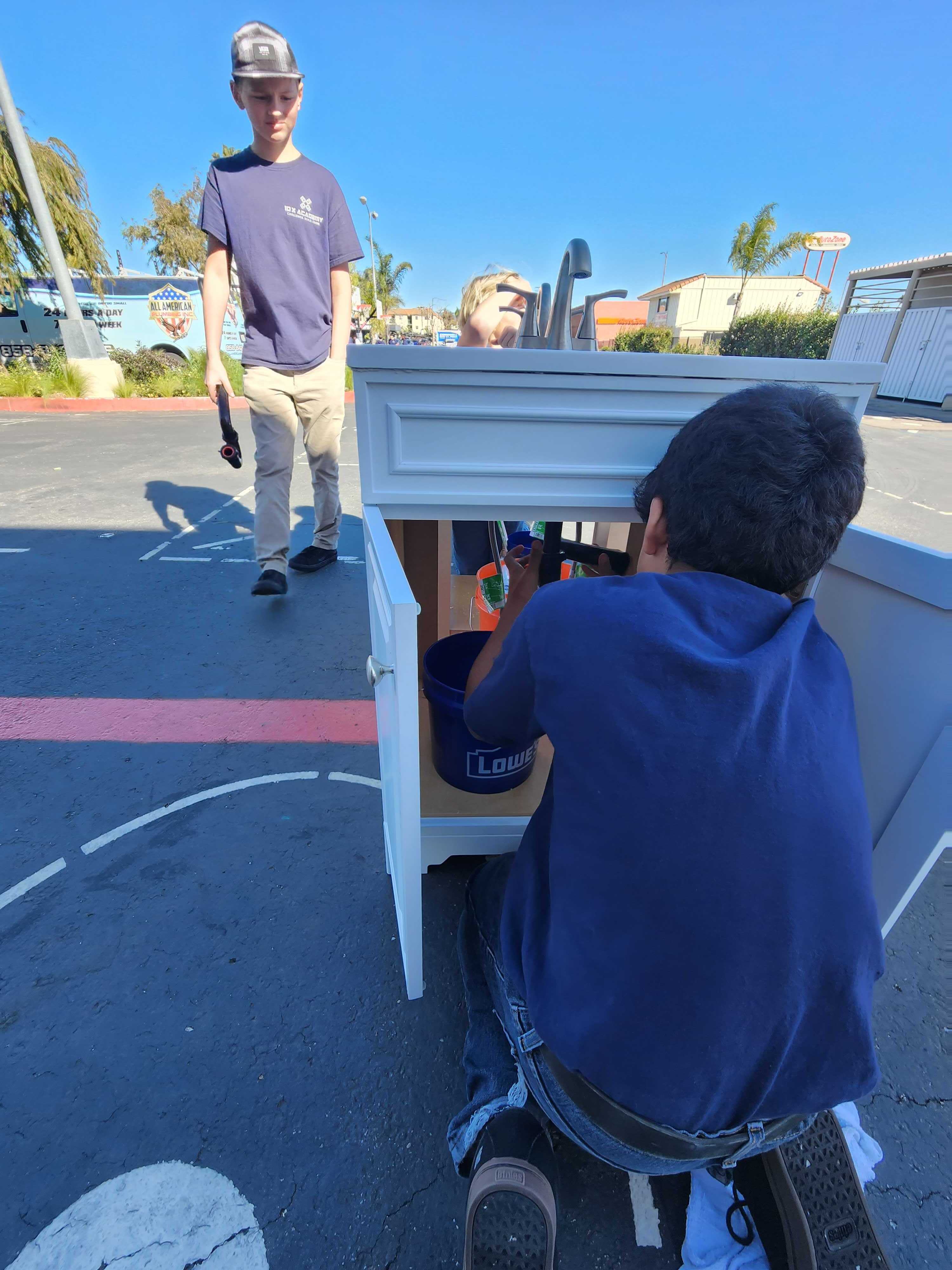 A boy kneeling installs plumbing under a white sink cabinet outdoors while another boy walks nearby holding a tool.