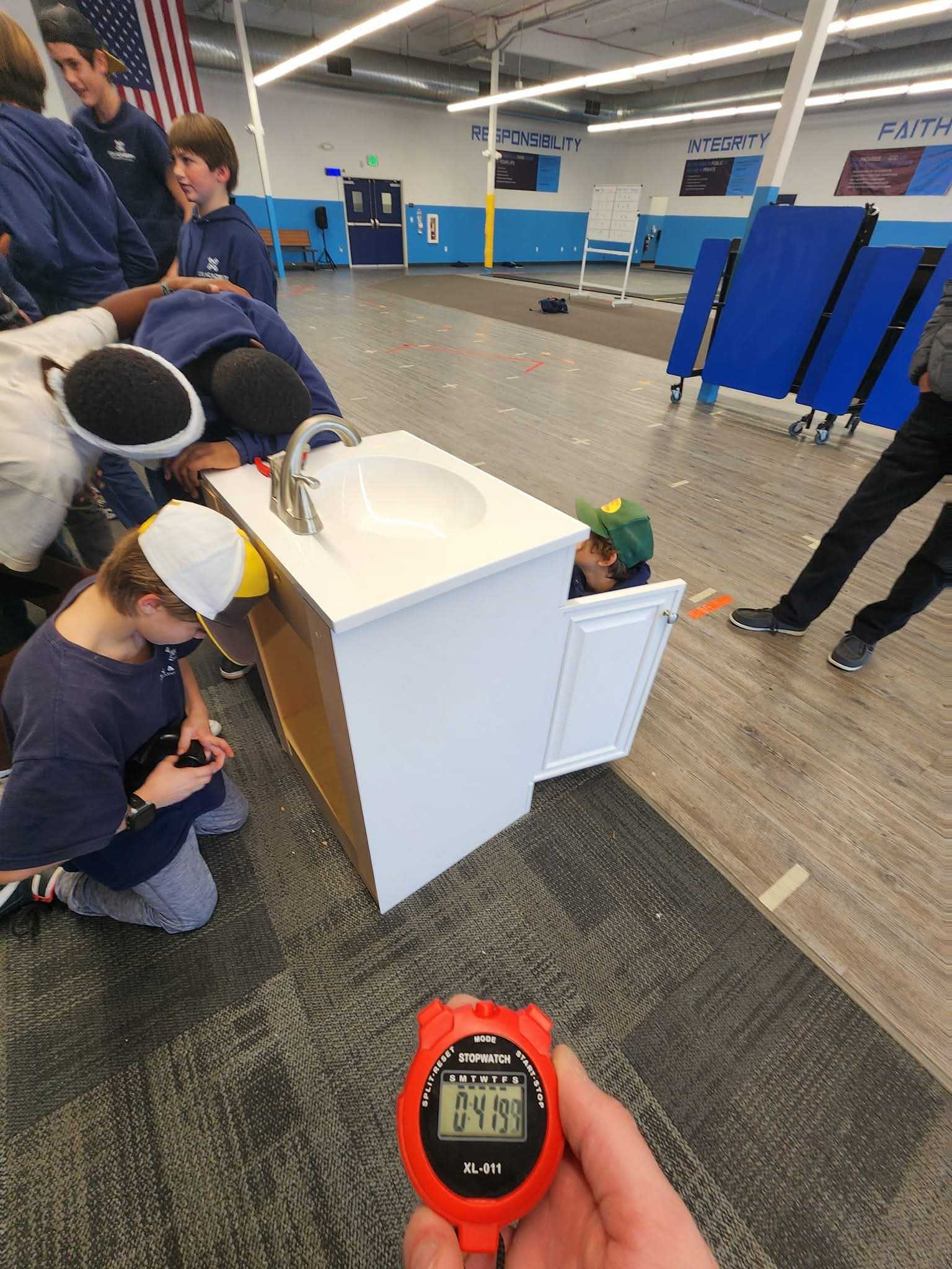 Several children gathered around and inside a cabinet with a sink in an indoor gym, with a hand holding a stopwatch showing 0:41.99 in the foreground.