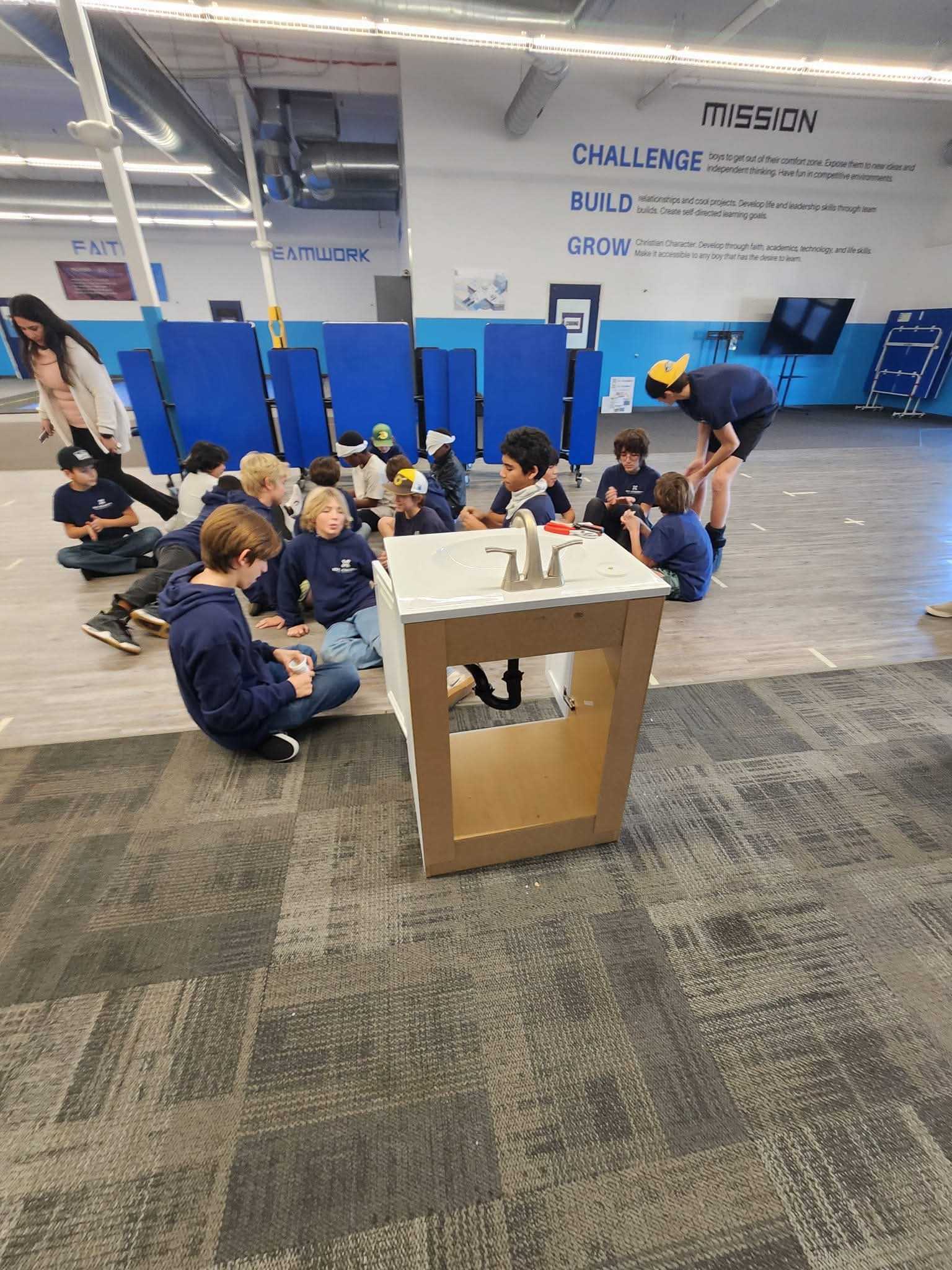 Group of children sitting on the floor in a room with a sink cabinet in the foreground and a mission statement on the wall behind them.