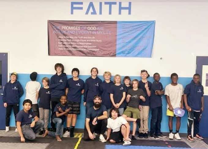 Group of diverse teenagers and one adult posing indoors under a banner and word 'FAITH' on the wall.
