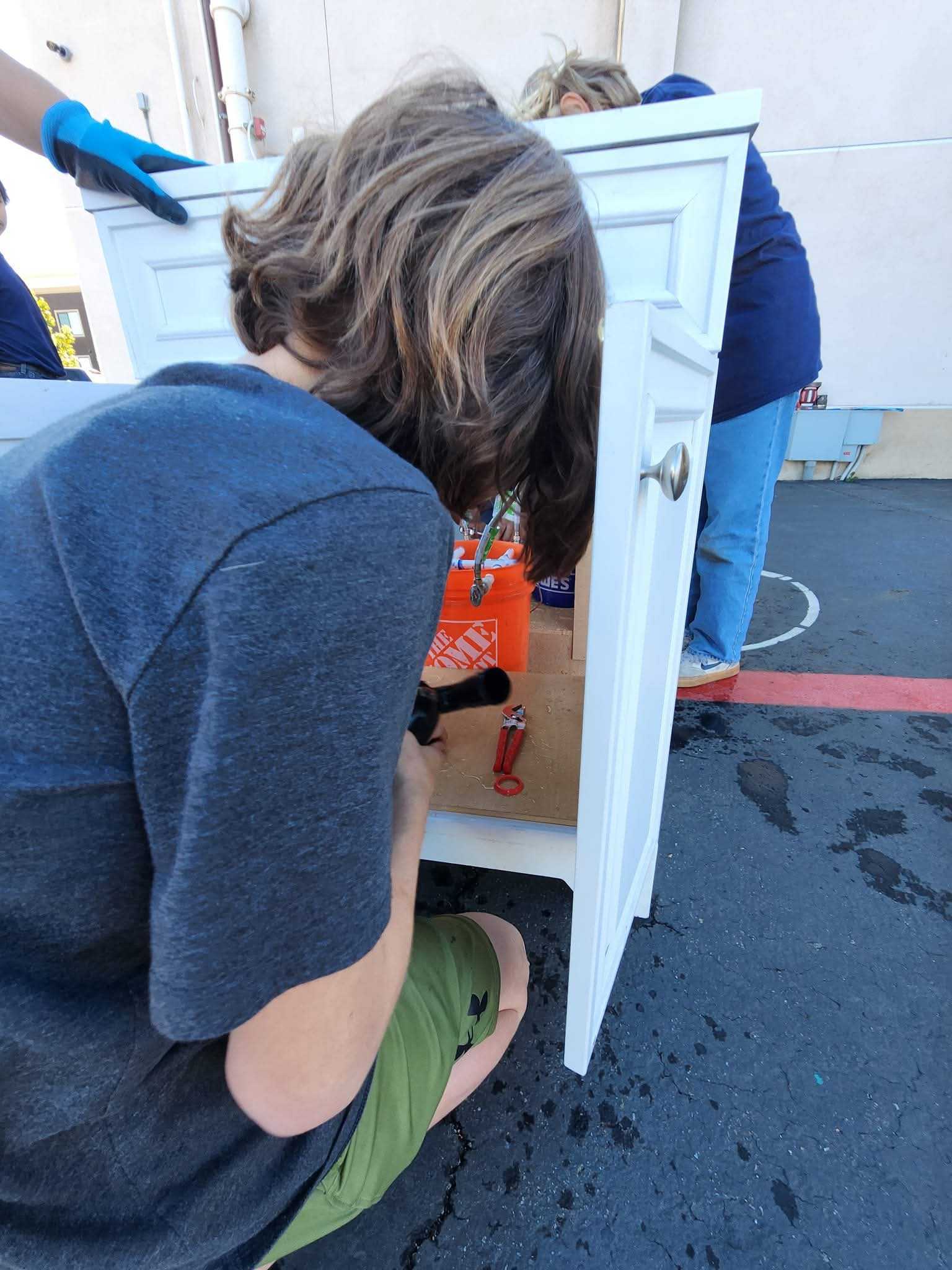 A person kneeling and working on plumbing inside an open white cabinet outdoors, with another person standing nearby.