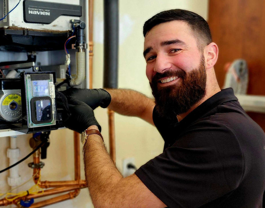 Man with a beard wearing black gloves and a black shirt working on a Navien heating system with copper pipes in the background.