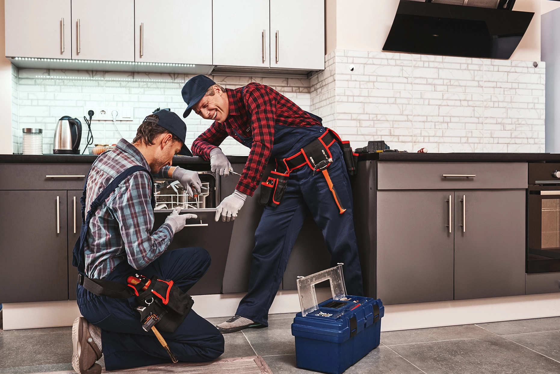 Two male plumbers in workwear repairing a dishwasher in a modern kitchen.